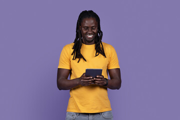 Joyful stylish young black guy using digital tablet on purple