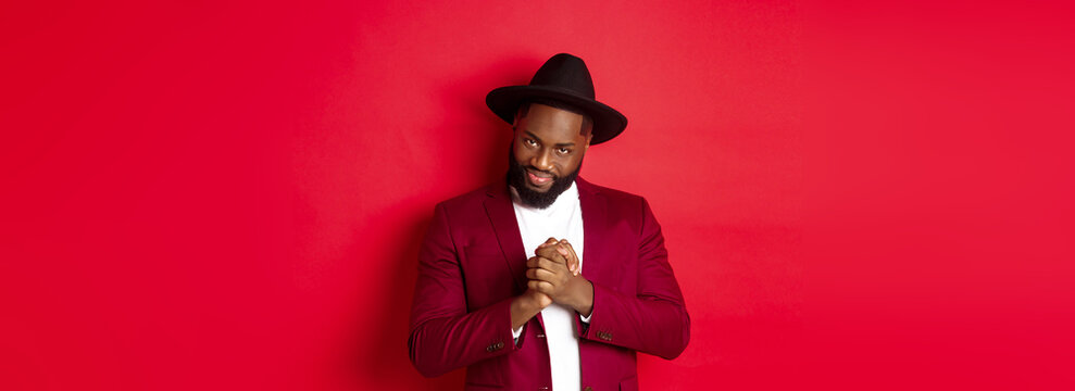 Cunning Black Man Having An Idea, Smiling Devious And Rubbing Hands, Standing Against Red Background And Scheming