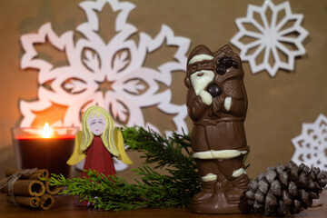 New Year's theme Santa Claus made of chocolate on a light brown blurred background with paper snowflakes with a Christmas tree branch and a cone with an angel figure and cinnamon and a burning candle