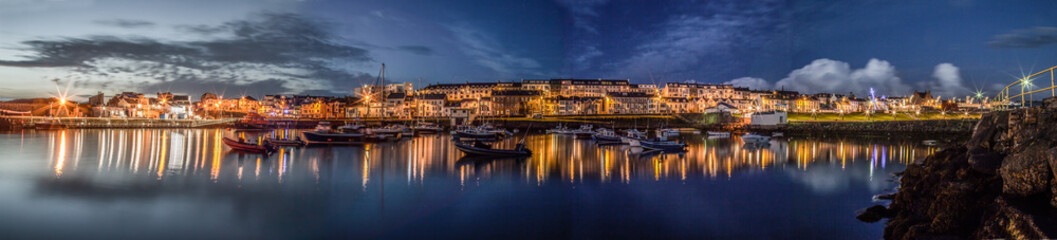 Fototapeta premium Portrush, Panorama of a small port town with a little harbour, boats and houses during a late summer night, blue hour, lights switched on