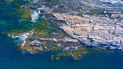 Looking down on rocky coastline with waves crashing and shallow water showing rocky layers...