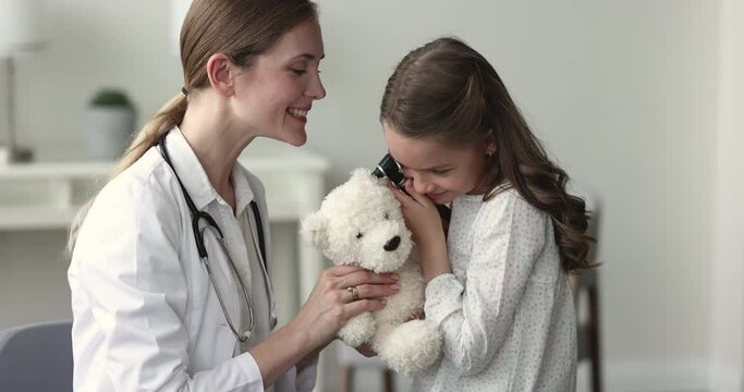 Little girl play ENT doctor hold auriscope check ear of fluffy toy bear during visit in clinic. Friendly paediatrician gives her professional medical tool to small patient to play during consultation
