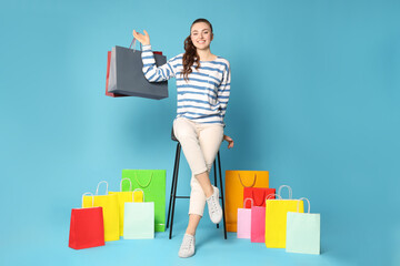 Happy woman holding colorful shopping bags on stool against light blue background