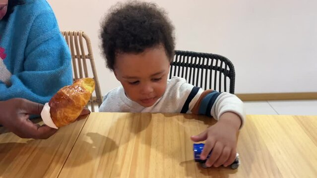Cute and exotic two-year-old black baby playing happy with his toy car inside a cafeteria while his mother feeds him with a croissant.