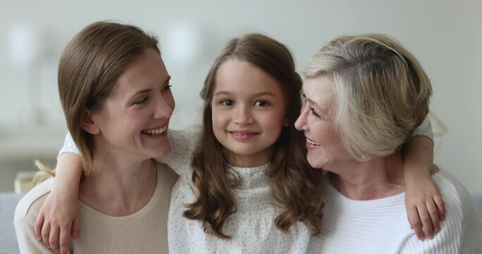 Close up portrait of three generational beautiful women smile look at camera, little girl hugging young mom and older granny sit indoors, feeling unconditional love appreciate each other. Family ties