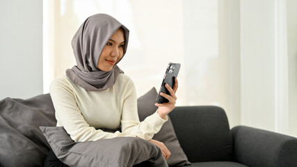 Beautiful Asian Muslim woman in hijab using her mobile phone while relaxing in her living room.