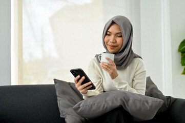 Charming Asian Muslim woman in hijab sipping coffee while using her mobile phone on the sofa