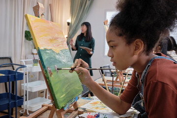African American girl concentrates on acrylic color picture painting on canvas with students group...