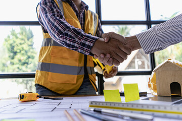 Engineers and construction crews shake hands, start new project contract plans in office center at construction site, partner and contractor in yellow helmets on table.
