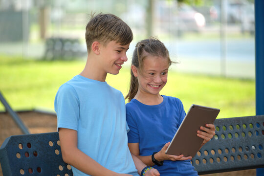 Two happy children, girl and boy looking at screen of digital tablet, reading, studying or gaming sitting on bench outdoors on summer sunny day