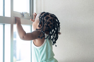 Cute African baby girl with her braids, playing near window, backside view of African pigtails for...