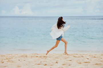 Sports woman runs along the beach in summer clothes on the sand in a yellow T-shirt and denim shorts white shirt flying hair ocean view
