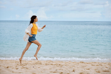 Fototapeta na wymiar Sports woman runs along the beach in summer clothes on the sand in a yellow tank top and denim shorts white shirt flying hair ocean view, beach vacation and travel