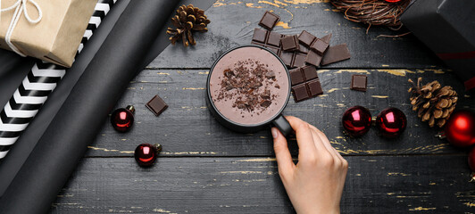 Female hand with cup of hot chocolate and Christmas balls on dark wooden background, top view