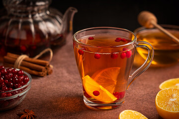Homemade healthy sweet hot tea drink served in transparent glass cup with red ripe cranberries and lemon citrus fruit slices on dark brown table with teapot, cinnamon spice and honey ingredients - Powered by Adobe