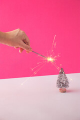 Christmas winter creative layout with a female hand holding a sprinkler and a small Christmas tree against a pink background. 80s or 90s retro aesthetic holiday concept. Minimal celebration idea