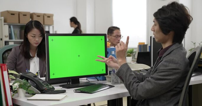 asian businessman using finger counting gestures while explaining project during a video call on computer with green screen in the office with coworkers