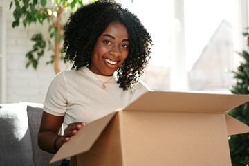 Young african woman sit on couch at home unpacking parcel cardboard box with online purchase