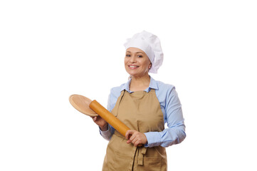 Beautiful Caucasian woman baker confectioner, pastry chef wearing beige apron, holding a rolling pin and wooden board, smiling a cheerful toothy smile, looking at camera, isolated on white background