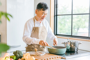 家のキッチンでフライパンを使って手料理を作るアジア人の高齢者男性（笑顔）
