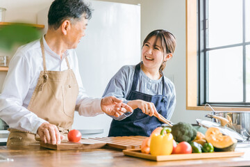 家のキッチンで料理・家事・自炊をして話す笑顔のアジア人高齢者男性と若い女性（家政婦・家事代行）
