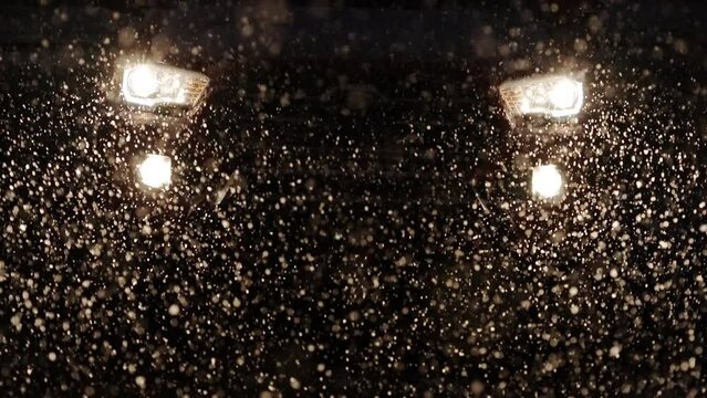 Snow falling at night in front of headlights from vehicle during storm as it moves in slow motion.