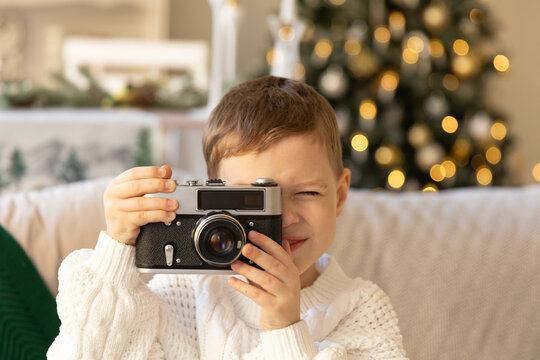 Adorable little child sitting on the sofa and playing with the retro photo camera in Christmas decorations. Baby boy taking picture with old camera. Indoors. Symbol of different generations.