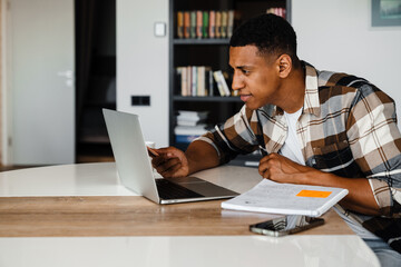 Young african man studying on laptop and writing down notes while sitting at table at home