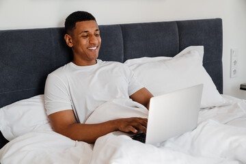 Joyful african man using laptop while lying in bed at home