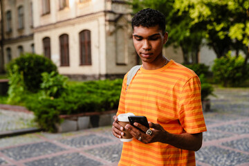 Young african man using smartphone and drinking coffee while standing outdoors