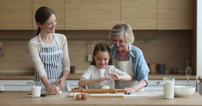 Little cute girl kneading and flattening dough preparing buns with help of caring young mother and aged grandmother cooking together in cozy kitchen at home. Home-made pastries, family recipe, cookery