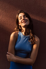 Beautiful young caucasian female looking at camera stands near building wall outside. Brunette wears blue top and leggins. Concept of city life, lifestyle.