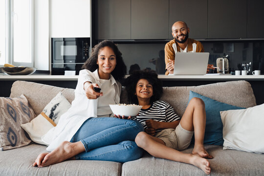 African American Mom And Son Eating Popcorn And Watching TV While Dad Working On Laptop