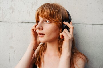 Ginger woman listening music with headphones while standing over gray wall