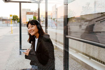 Fototapeta premium Young asian woman using cellphone and headphones sitting on bus station