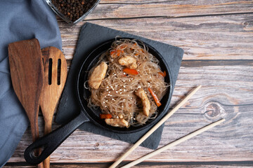 Fried funchosa in a frying pan with chicken on a wooden background.