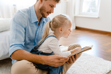 Young white father and little daughter reading book together at home
