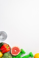 Proper nutrition concept. Top view vertical photo of plate with vegetables fruits bell pepper cauliflower cucumber grapefruit glass of water and dumbbells on isolated white background with copyspace