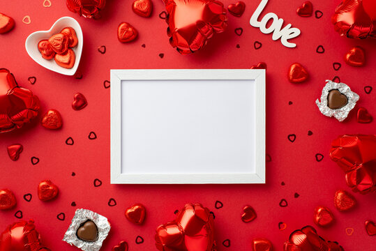 Valentine's Day concept. Top view photo of white photo frame heart shaped balloons chocolate candies plate inscription love and confetti on isolated red background with copyspace