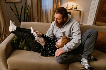 Father and little son reading book while resting on sofa together