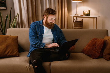 Smiling redhead man using laptop computer while sitting on sofa in living room