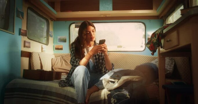Young Beautiful Digital Nomad Relaxing in a Caravan, Using Smartphone to Check Social Media Updates, Message Friends and Make Some Online Shopping. Female on a Camping Trip in a Motorhome.