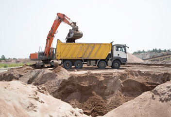 Obraz premium orange crawler excavator pours earth into body of yellow dump truck. Working moment at construction site in process of earthworks. Loading and transporting soil on construction vehicle.