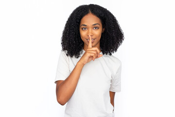 Pleased multiracial woman pressing finger to lips. Portrait of happy young female model with dark curly hair in white T-shirt looking at camera, smiling and shushing. Silence, secret concept