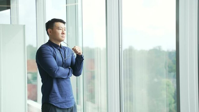 Successful thoughtful IT specialist businessman employee looking at window in modern office centre Confident asian entrepreneur professional developer pensive thinking about career deal alone indoors