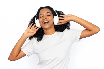 Happy multiracial woman dancing in headphones. Excited young female model with dark curly hair in white T-shirt with closed eyes, smiling, enjoying music. Modern technology, music concept
