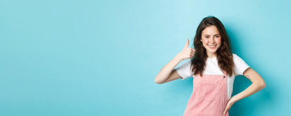 Portrait of confident and positive young woman show thumb up, say yes, give permission, approve and agree something good, praise nice choice, standing against blue background - obrazy, fototapety, plakaty