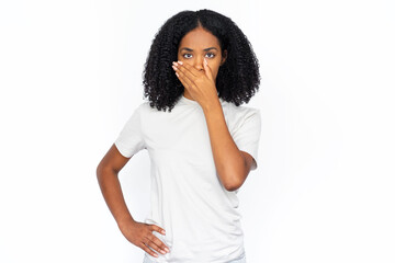 Serious African American woman covering mouth. Portrait of surprised young female model with dark curly hair in white T-shirt looking at camera, asking to be quiet. Silence, shock concept