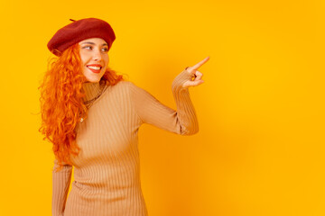 Red-haired woman in a red beret on a yellow background, studio shot, pointing at copy space
