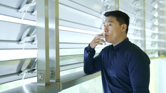 Confident asian businessman smoking cigarette during break at workplace alone Portrait of attractive entrepreneur man in blue shirt relaxing standing indoors modern office buildings Business lifestyle